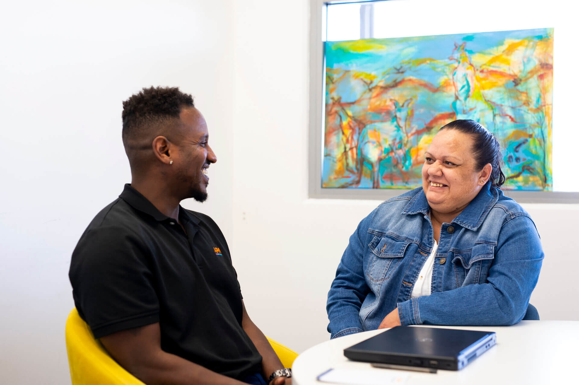 A man in a black APM polo sits at a table with a smiling Indigenous woman