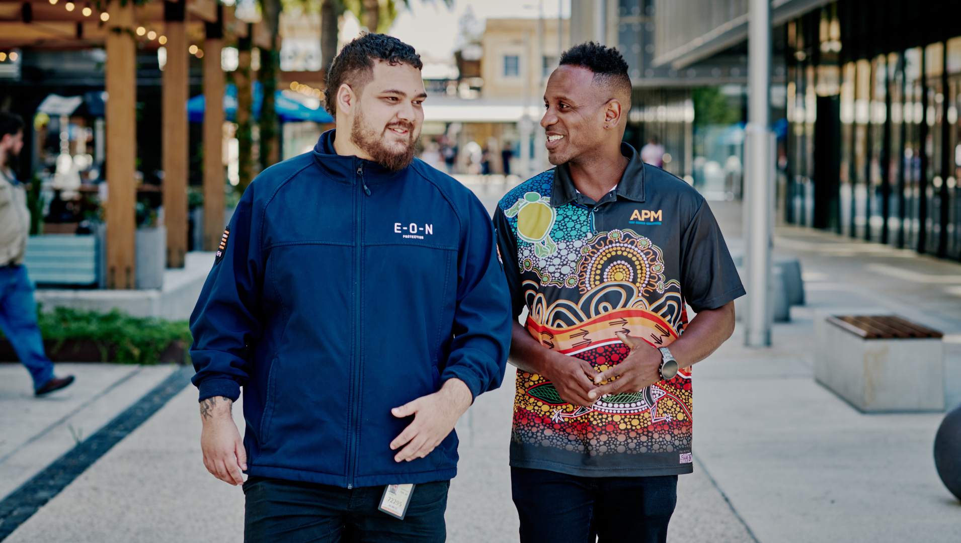 A man in blue jacket smiles with an APM consultant in an Indigenous polo